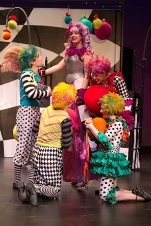 The fairies in fancy wigs, black and white silks and colorful skirts surround purple-haired Titania with a breastplate armor and balloons and the checkerboard backdrop in the background